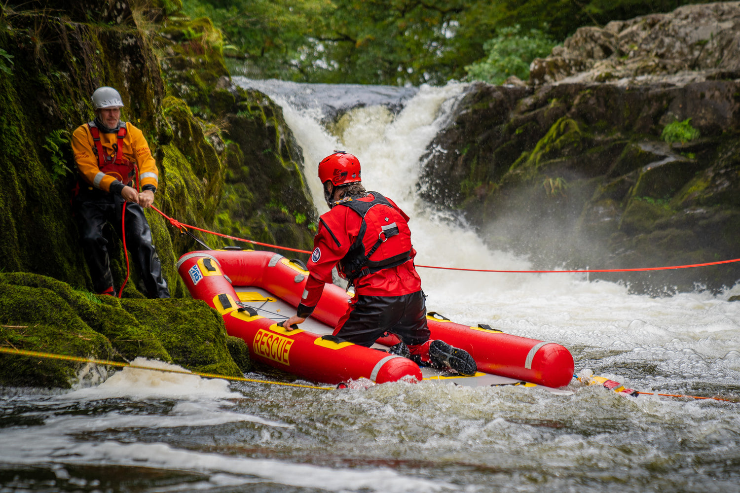 A red rescue raft with two people in a river setting near a waterfall, with another person standing on a mossy rock holding ropes. The raft has yellow and black accents and the word 'RESCUE' printed on it.