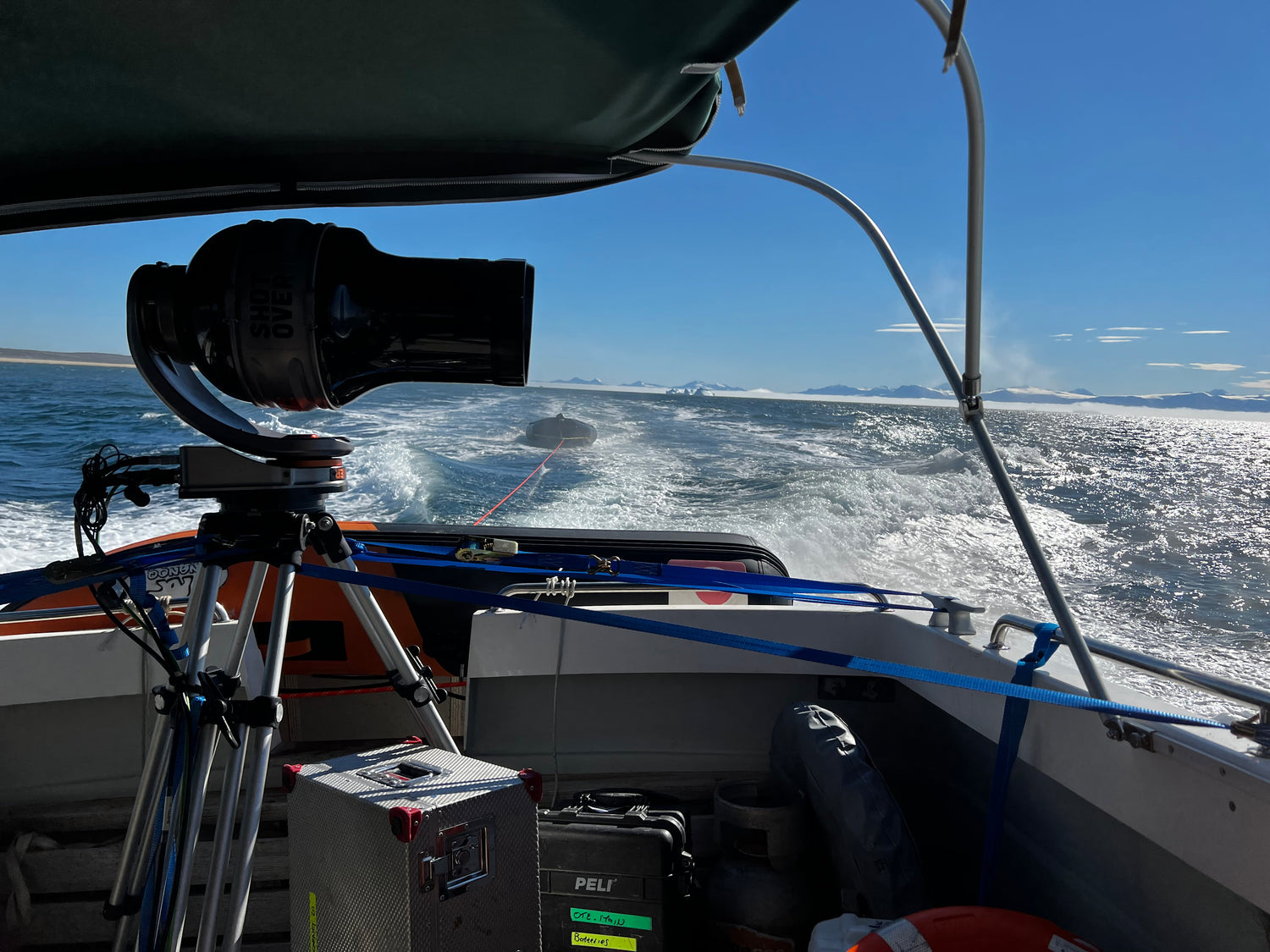 A camera mounted on a tripod aboard a moving boat, with ocean water and distant land visible under a blue sky. The boat interior, rigging, and equipment are visible in the foreground.
