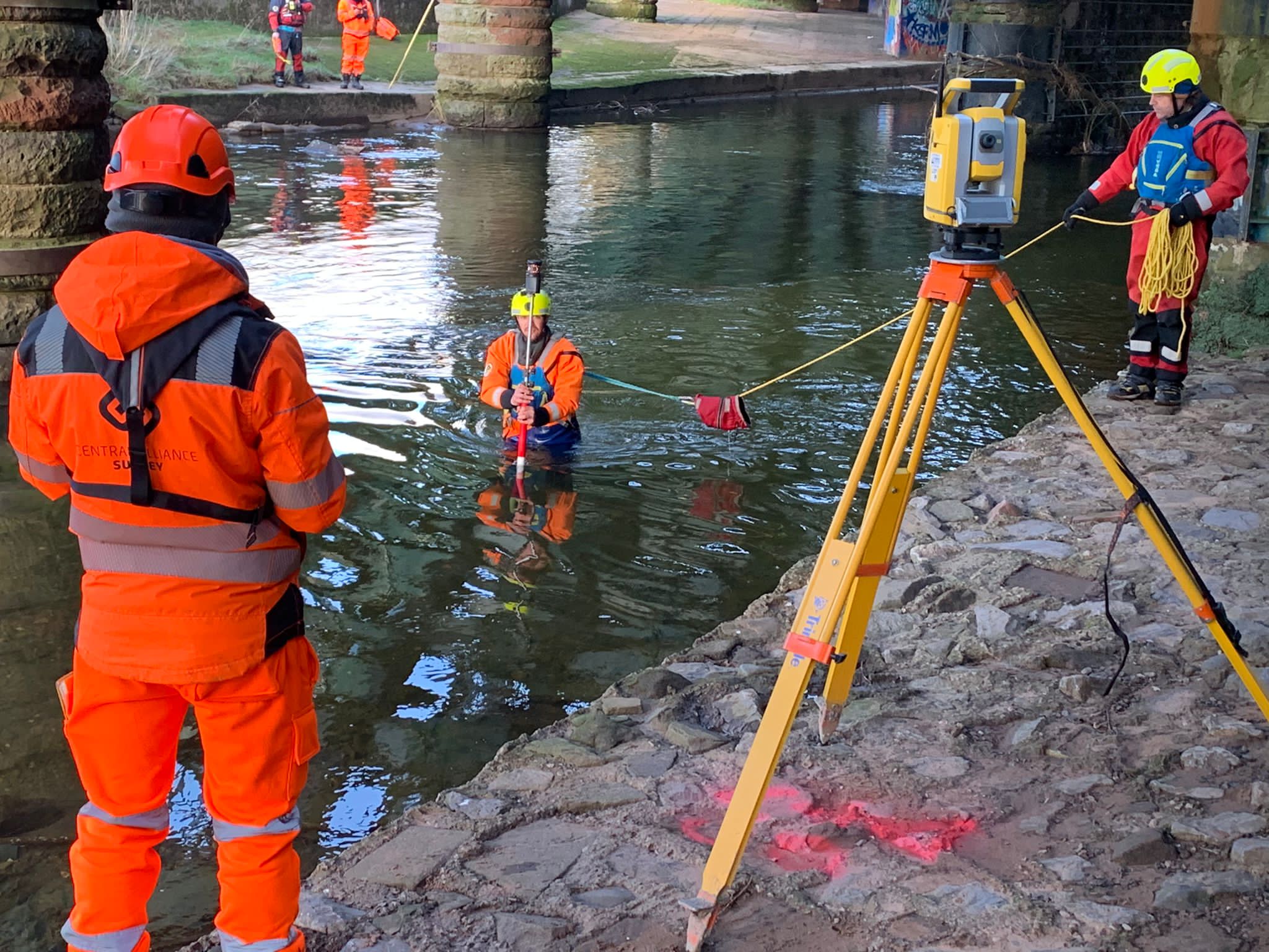 Workers in high-visibility orange safety suits and helmets conduct a survey or measurement operation in shallow water, with a yellow tripod-mounted surveying instrument on the shore and another worker standing nearby.