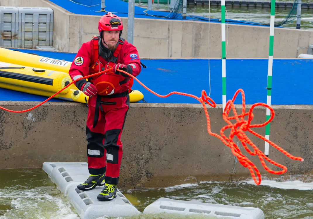 A rescue worker in a red and black uniform with a red helmet stands on a concrete platform in shallow water, holding an orange rope. A yellow inflatable raft is visible in the background next to a blue padded area.