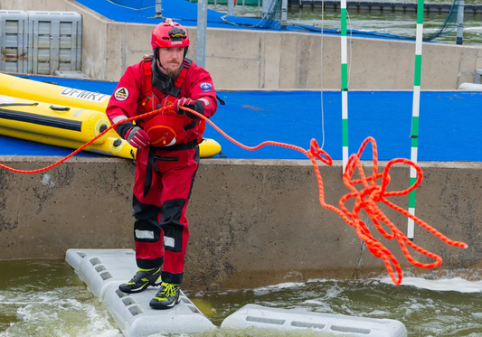 A rescue worker in a red and black uniform with a red helmet stands on a concrete platform in shallow water, holding an orange rope. A yellow inflatable raft is visible in the background next to a blue padded area.